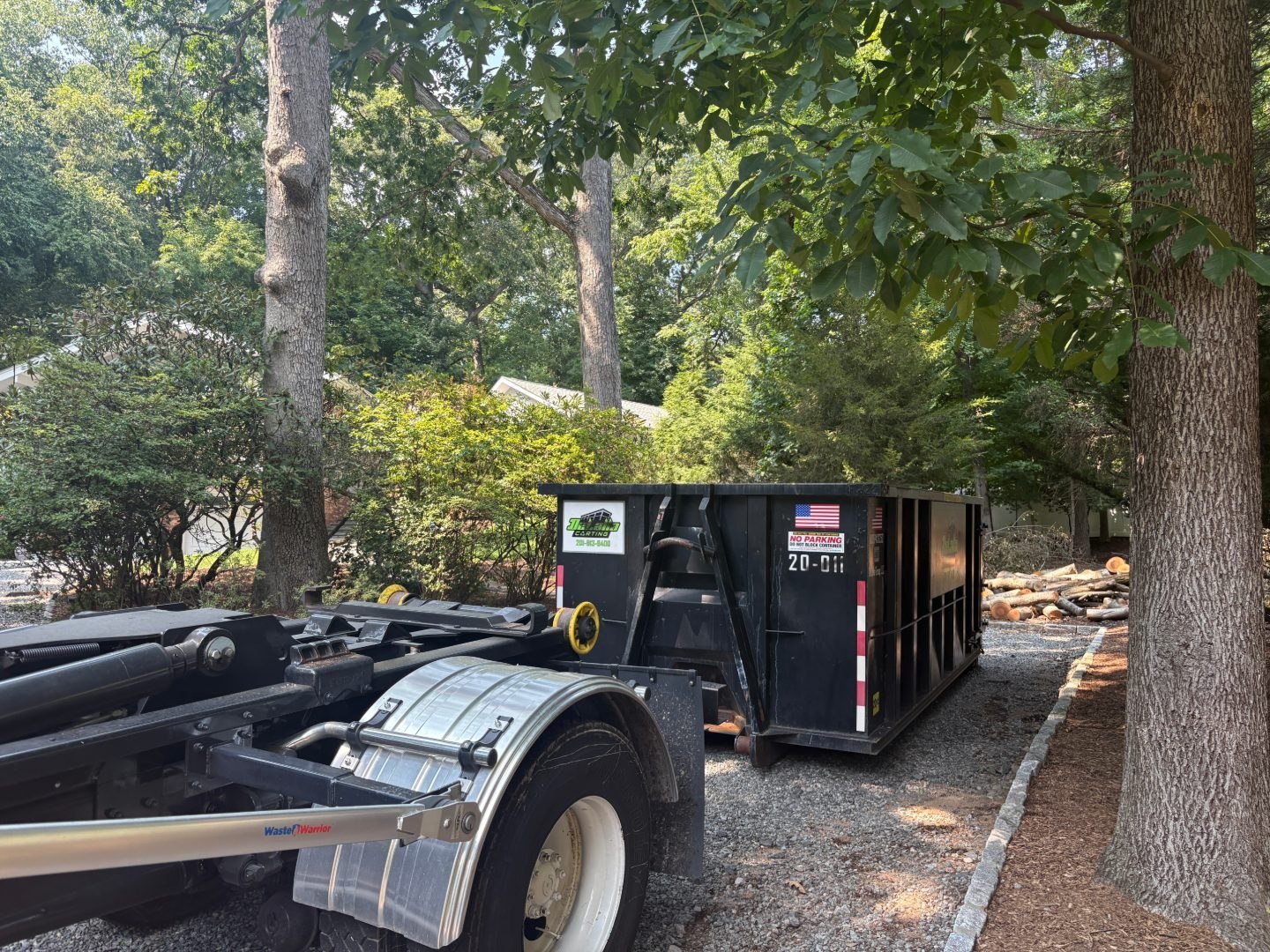 20-yard dumpster on a gravel driveway in a wooded lot in Paramus, NJ, cut logs stacked for loading.