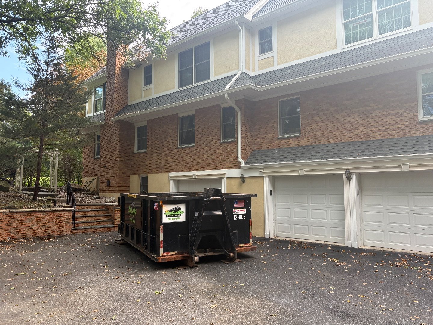Black roll-off dumpster parked on a driveway beside a brick house with white double garage doors and trees.