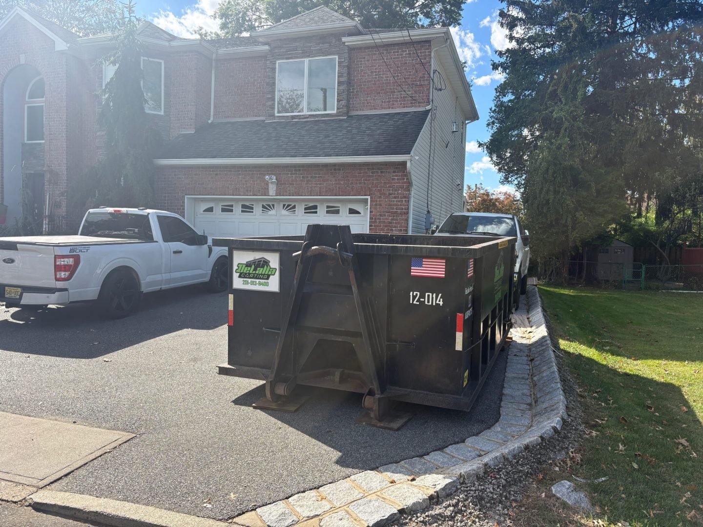 Large black dumpster on a paved residential driveway beside two white pickup trucks and a brick house.