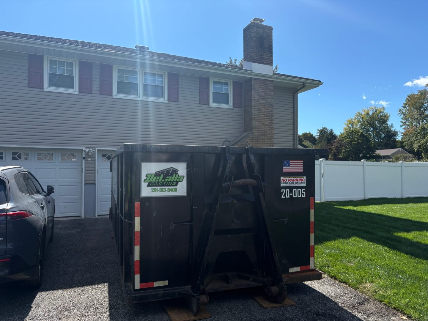 Black roll-off dumpster on boards in front of a two-story house and garage, with a parked SUV to the left.