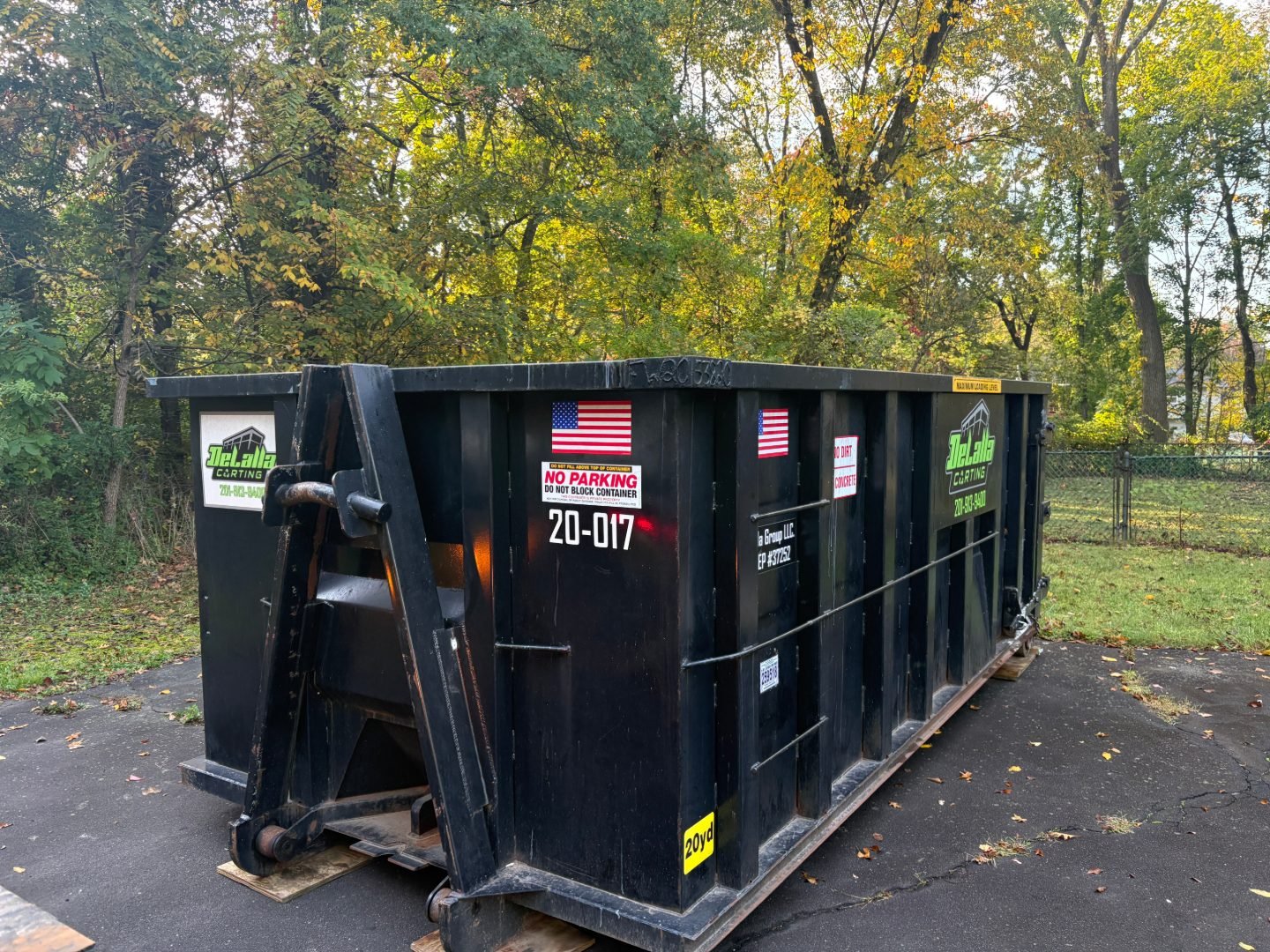 Large black roll-off dumpster with decals and warning signs on a paved driveway surrounded by leafy trees.