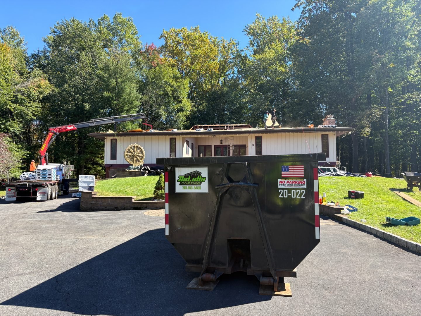 Black dumpster on a driveway beside a crane truck and a house with workers repairing the roof.
