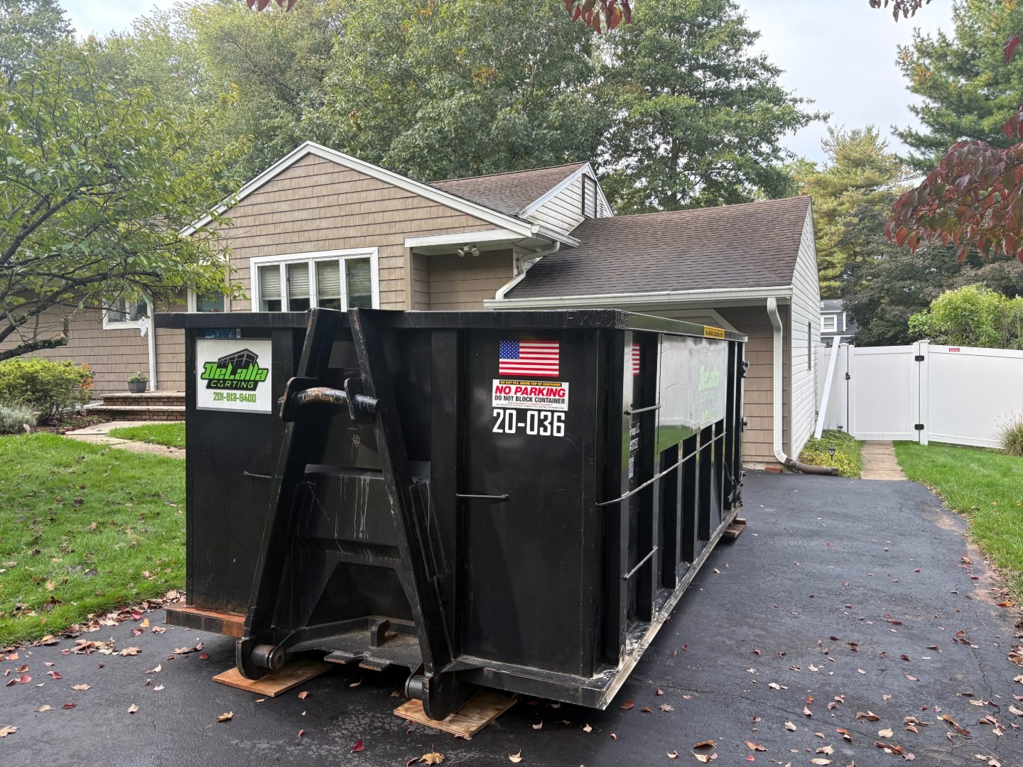 Large black roll-off dumpster placed on boards in a residential driveway beside a house and white fence.