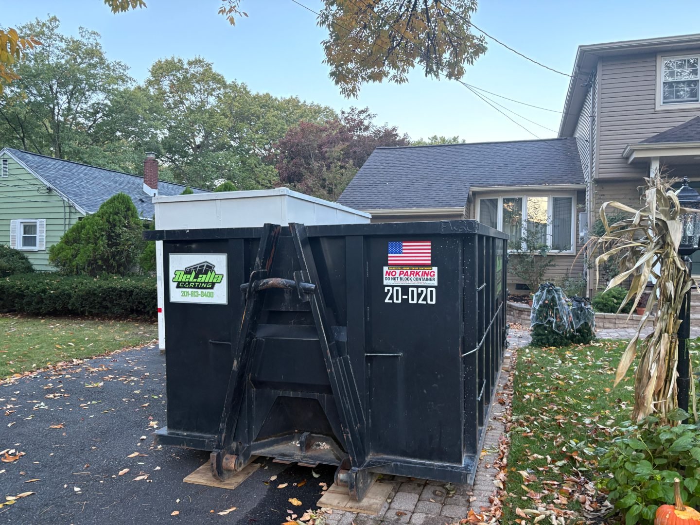 Black roll-off dumpster placed on boards in a residential driveway with fallen leaves and autumn decorations nearby.