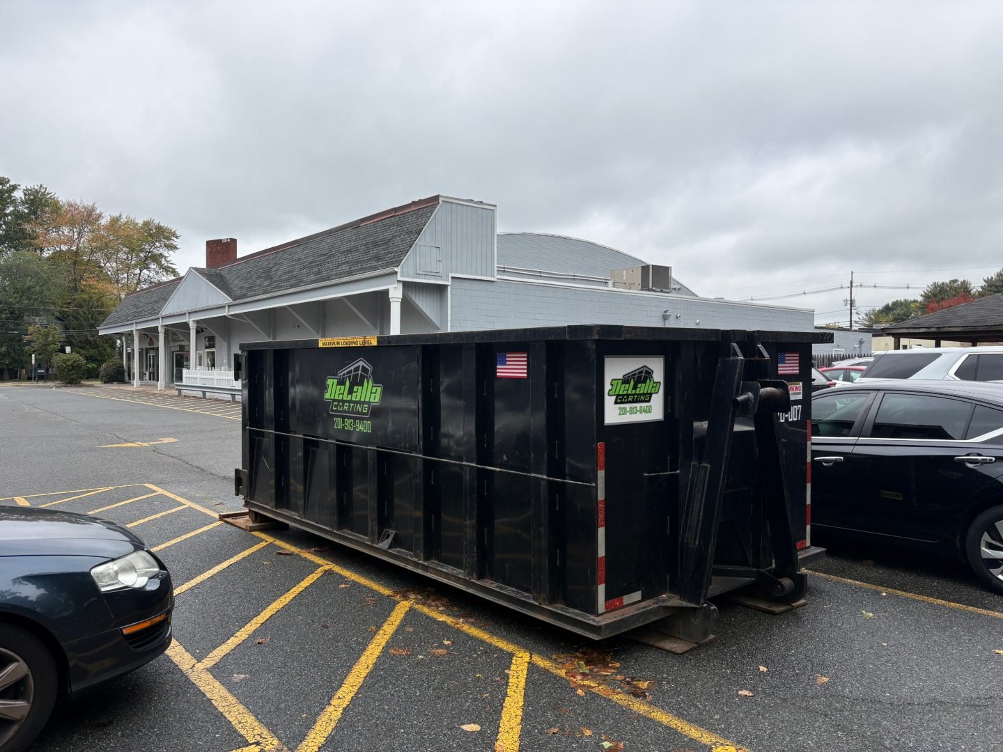 Large black roll-off dumpster in a yellow-striped parking area beside parked cars and a gray building.