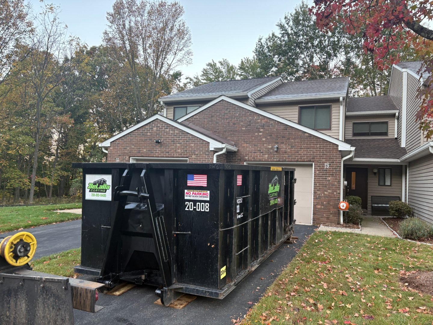 Black roll-off dumpster positioned in a residential driveway in front of a brick house, with autumn trees and fallen leaves.